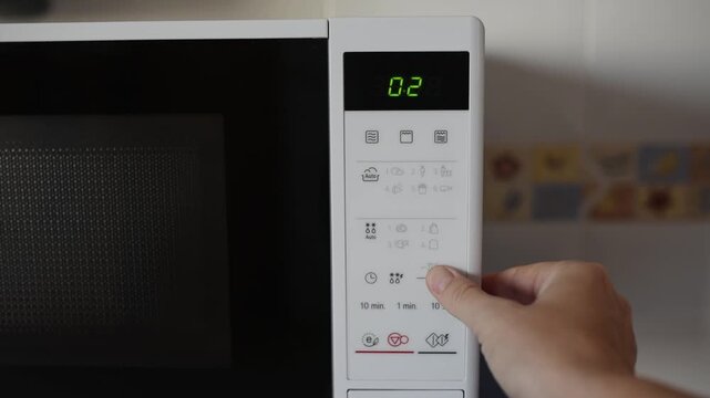 User hand selects defrost mode and sets time on digital display of microwave. Video illustrates use of household appliances, fast food preparation, home routine and modern kitchen technologies.