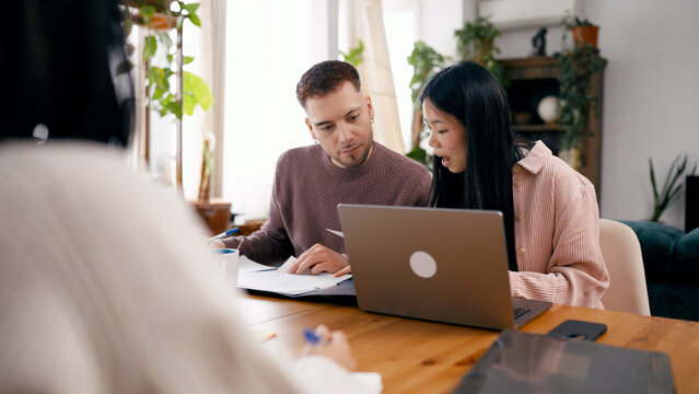Couple reviewing documents and using a laptop while meeting with a financial advisor at a wooden table in their home