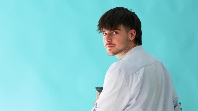 young caucasian male student with back to camera turns to face viewer, smiles confidently, then looks down at smartphone, isolated on blue background