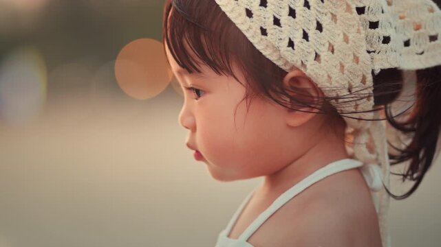 Side view of a cute Asian toddler girl wearing a white crochet bandana, gazing at the sea during a golden hour sunset at the beach. A peaceful moment of childhood wonder and summer vacation. Cinematic