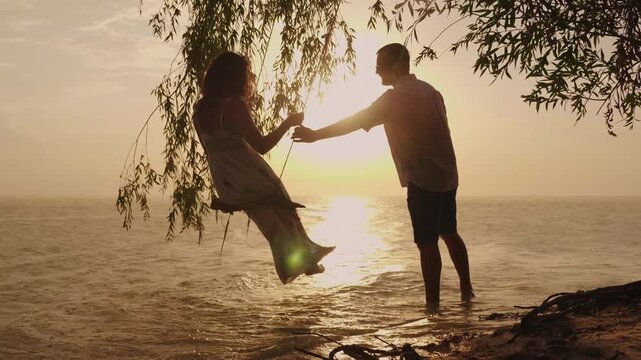 Silhouette of man pushing woman on rope swing at sunset beach during summer vacation.
