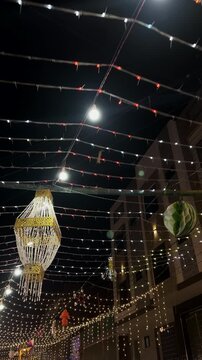 Low angle shot moving beneath strings of fairy lights, decorative lanterns, paper ornaments, and festoon bulbs hanging above the center of a street at night during 12 Rabi ul Awal in Pakistan.