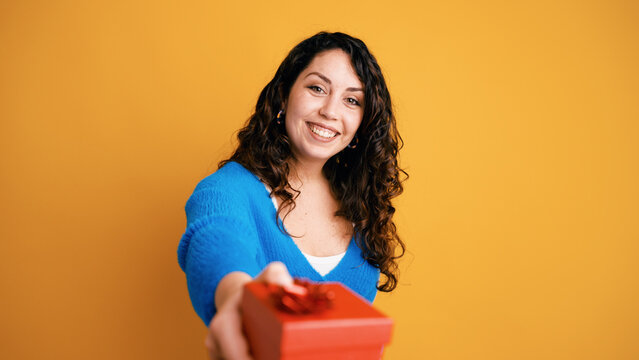 Happy young woman extending a red present box, sharing joy and generosity on a yellow background, celebrating special moments