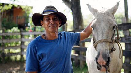 Mature Latino ranch worker in hat stands by white horse at sunny farm fence, smiling confidentlyexperienced equestrian lifestyle portrait © DMegias