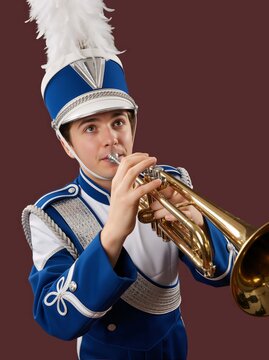 Young male marching band musician playing a brass trumpet. High school student in blue and white uniform with feathered shako hat. Studio portrait on solid maroon background