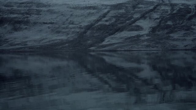 Serene dark water reflects textured rocky shore under moody sky