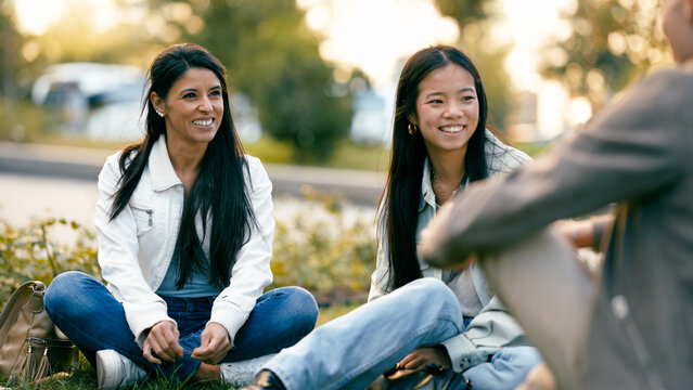 Smiling diverse young women sitting on grass in a park, casually talking, laughing and enjoying time together outdoors on a sunny day