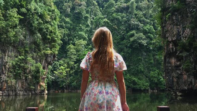 Young woman in floral dress looking at tropical lagoon surrounded by lush limestone cliffs and dense jungle during daytime. Back view of traveler exploring scenic nature in Southeast Asia.