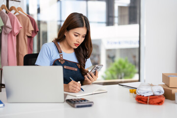 Fototapeta premium Small business owner wearing an apron, jotting down notes on a notepad while working on cell phone.
