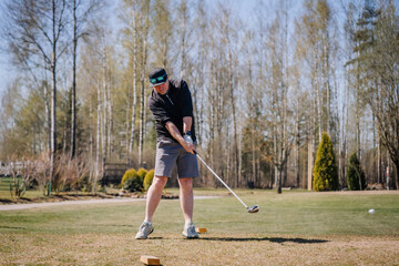 Fototapeta premium Male golfer swings club on a sunny day at a golf course with trees and grass in the background