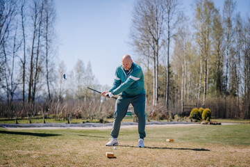 Obraz premium Senior man in teal athletic wear swings golf club on sunny day at outdoor golf course with trees and grass in the background