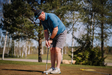 Obraz premium Senior man putting golf ball on green during sunny day, surrounded by trees and open space on a golf course