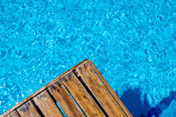 Wooden pool deck beside bright turquoise swimming pool water with shimmering reflections and rippling surface textures creating a calm summer leisure background for travel resort and relaxation themes © Duncan
