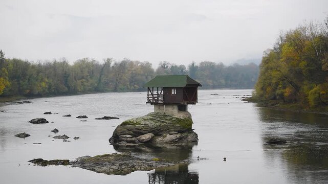 Lonely cottage on rock in middle of Drina river near Bajina Basta in Serbia, autumn colors on forest on riverbanks and fog obscures mountains in background, popular tourist attraction