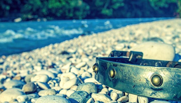 Ancient style steel neck gorget with rounded edges and decorative brass fasteners resting on a pile of smooth river stones by a flowing water bank, captured in a cinematic widescreen composition.