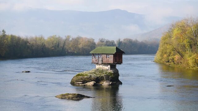 Lonely cottage on rock in middle of Drina river near Bajina Basta in Serbia, autumn colors on forest on riverbanks and mountains in background are in haze, popular tourist attraction
