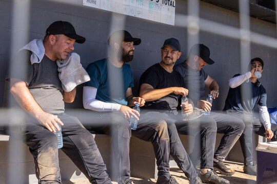 Diverse male teammates sitting on wooden bench in dugout, drinking from clear plastic bottles