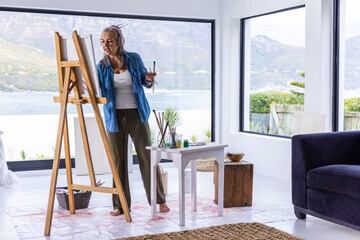 Naklejka premium Senior female painter standing at easel, painting on canvas in bright studio wearing headphones