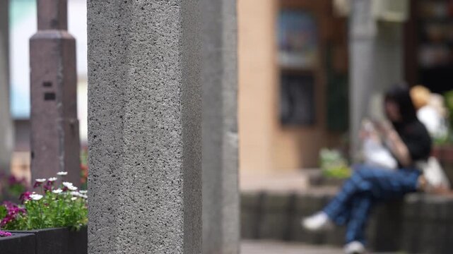 Granite pillars frame a quiet street scene. Person sits, absorbed in their phone. Another walks past, unaware of the moment. Flowers bloom softly beside stone. Urban calmness captured in shallow depth