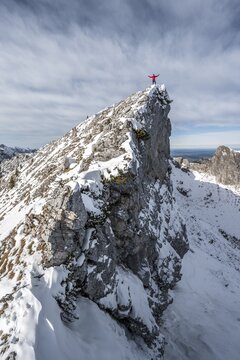 Mountaineer stretching his arms in the air, standing on a snow-covered rocky ridge, Fensterl on the hiking trail to Ammergauer Hochplatte, in autumn, Ammergau Alps, Bavaria, Germany