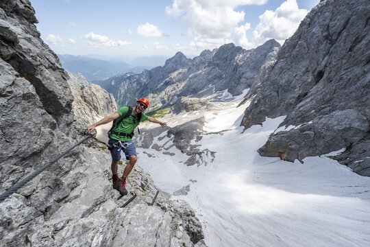 Mountaineer with helmet in a secured via ferrata, Zugspitze via ferrata, ascent to the Zugspitze, view of mountain basin with snow field of the H&ouml;llentalferner and H&ouml;llental, Wetterstein range, Bavaria, Germany