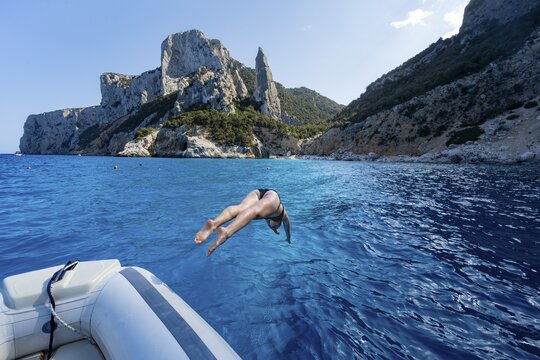 Young woman jumping from a boat into the water, picturesque rocky coast, cliffs with L'Aguglia rock pin, blue sea and Cala Goloritz&eacute; beach, Golfo di Orosei, Baunei, Sardinia, Italy