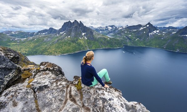 Young hiker sitting on cliff, precipice to fjord, view from Mount Barden, Senja, Norway