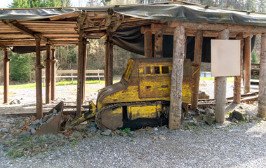 Fototapeta premium Weathered yellow bulldozer with front blade and crawler tracks stands under a rough wooden shelter with rocks around it. The scene shows abandoned construction machinery, quarry work, industrial decay