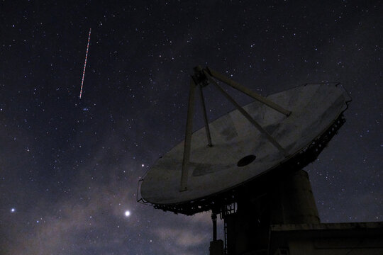Radio telescope antenna under starry night sky with light trail