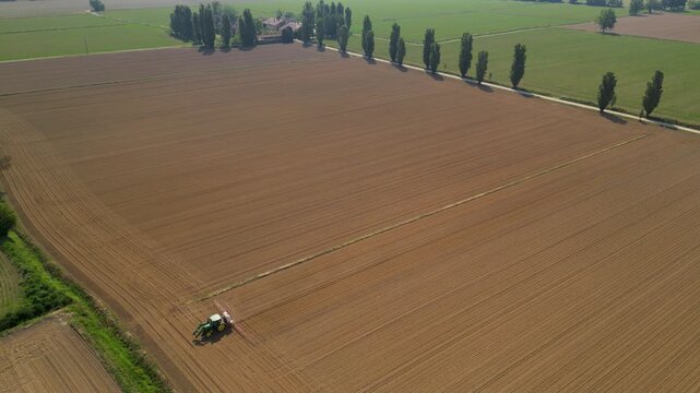 Green tractor cultivating a large, freshly tilled brown field, with a row of tall trees lining the edge under a clear sky.