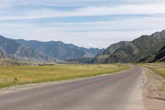 Empty asphalt road of the Chuysky Trakt running through a wide mountain valley with green meadows and dramatic ridges, Altai Republic, Russia, sunny day