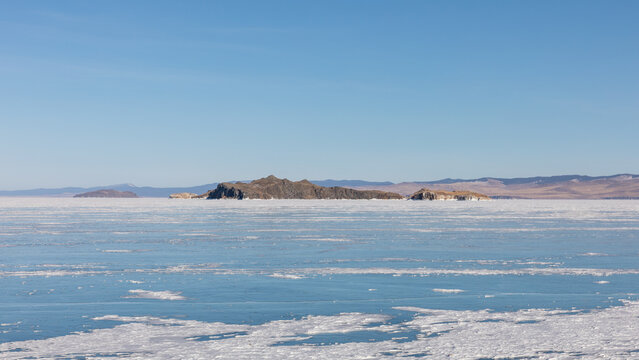 Wide winter view of frozen Lake Baikal with blue ice, cracks and scattered snow, distant rocky islands and clear sky in Maloye More, Irkutsk Oblast, Russia