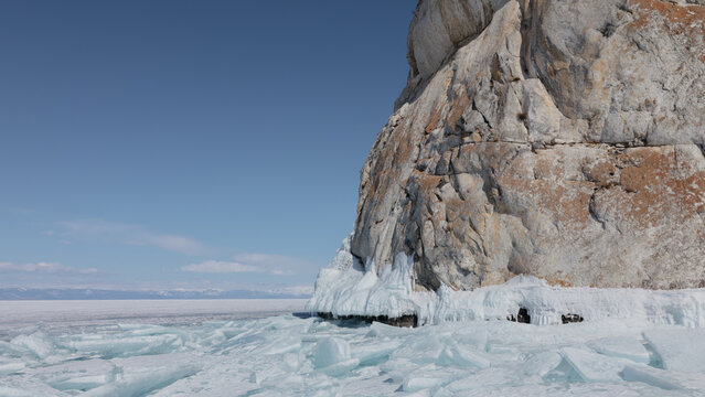 Rugged rock cliff with blue ice formations and broken ice hummocks on the frozen surface of Lake Baikal in winter, Siberia, Russia, under clear sky