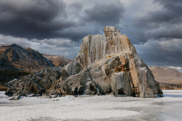 Massive granite rock formation rises above the frozen surface of Lake Baikal in Siberia, Russia, with dark storm clouds and rugged mountains creating a moody winter landscape © TravelFlow