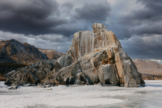 Massive granite rock formation rises above the frozen surface of Lake Baikal in Siberia, Russia, with dark storm clouds and rugged mountains creating a moody winter landscape