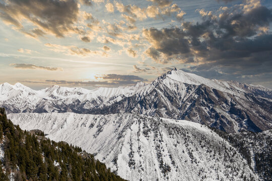 Wide panoramic landscape of Pik Lyubvi above Arshan village, Buryatia, Russia, with snow-covered ridges, dark conifer forest and dramatic clouds at sunset