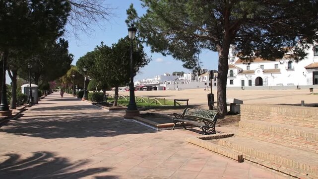Peaceful tiled walkway with trees, benches, and traditional white Andalusian architecture under a clear blue sky in El Rocio.