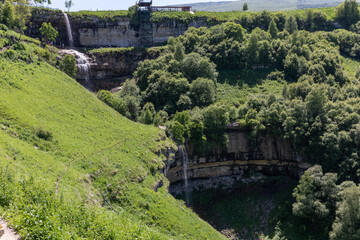 Fototapeta premium Scenic view of Matlas waterfalls flowing over layered limestone cliffs into a lush green gorge with trees and hiking viewpoint, Dagestan, Russia, sunny day
