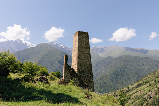 Stone watchtower ruins of the Erzi tower complex on a green mountain slope with snowy Caucasus peaks and blue sky, Ingushetia, Russia