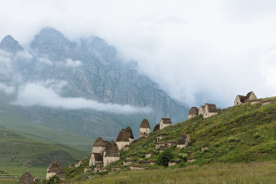Scenic view of the Dargavs City of the Dead necropolis with ancient stone tombs on a green hillside, misty mountains behind, North Ossetia&ndash;Alania, Russia