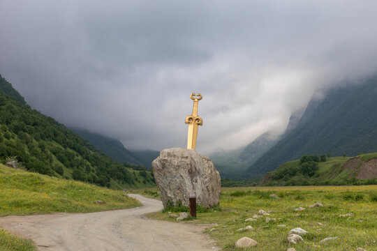 Golden sword sculpture embedded in a boulder beside a dirt road in the Midagrabin Valley, North Ossetia, Russia, with green mountains and low clouds