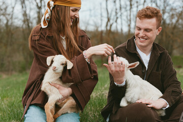 Fototapeta premium Cheerful couple outdoors with a goat kid, gentle interaction in a sunlit meadow, rustic vibe, warm colors, natural lighting, candid portrait of bonding and playful affection with animals