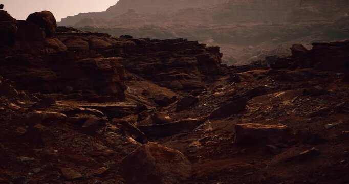 A wide rugged canyon of red rock ledges and scattered boulders descends toward hazy distant ridges under soft warm light.