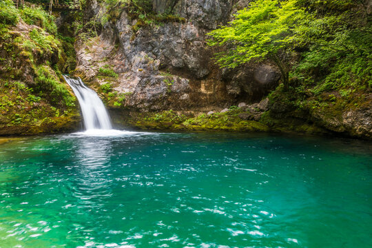 Wide shot capturing the emerald-blue waters of the Blue Eye of Theth, with a waterfall flowing into a vibrant pool framed by rugged mountains in Theth, Albania.