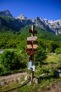 A multi-directional wooden signpost on a grassy trail, featuring labels, set against a stunning backdrop of lush green forests and jagged mountain peaks in Valbona Valley, Albania.