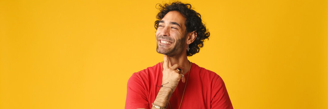 Pensive attractive man with curly hair, dressed in red T-shirt,  thinking something, having good idea isolated on yellow background in studio. Panoramic