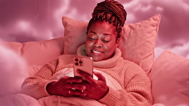 Woman Relaxing with Funny Videos on Phone - An African-American woman is lying in bed surrounded by pillows and blankets, watching videos on her phone.