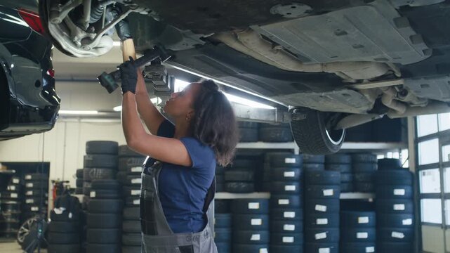 Focused female mechanic using a torch for visibility, inspecting the vehicle's underside in a professional automotive workshop.