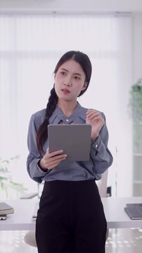 young Asian businesswoman professional using digital tablet computer standing thinking reading business report data analysis at modern bright office workspace