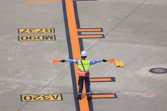 Back view of an aircraft marshaller signaling with orange paddles to guide a passenger plane to its designated parking position on the airport apron.